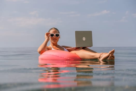 A woman is floating on a pink inflatable raft in the ocean while holding a laptop. Concept of relaxation and leisure, as the woman is enjoying her time in the water while working on her laptop