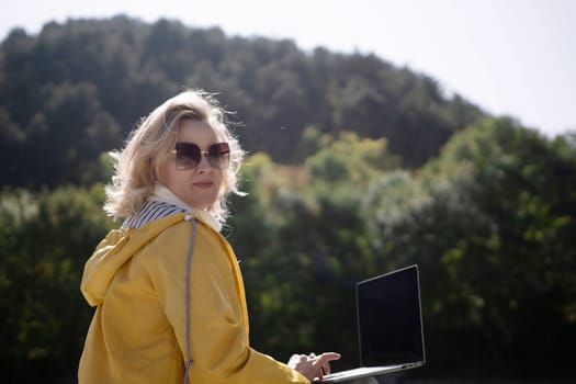 A woman in a yellow jacket is sitting on a bench with a laptop in front of her. She is smiling and she is enjoying her time outdoors