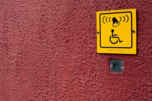 Wheelchair sign with braille text next to staff assistance call button on red structural wall, handicapped access symbol