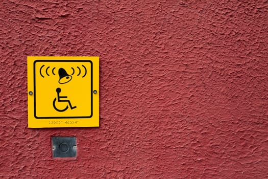 Yellow wheelchair icon with bell on red wall next to button to call help for disabled person, special button and sign for people in wheelchair with braille text, accessible environment for the disabled