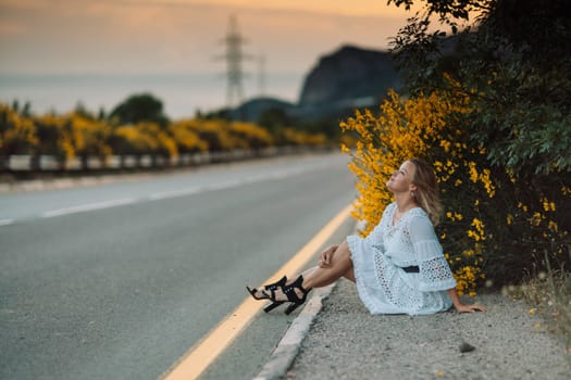 A woman is sitting on the side of a road, wearing a white dress and black shoes. The scene is peaceful and serene, with the woman looking out at the road and the surrounding landscape
