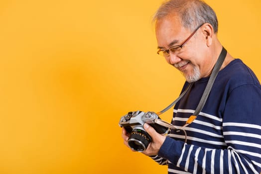 Portrait Asian old man wearing glasses look picture on screen vintage SLR camera studio shot isolated yellow background, smiling happy Photographer elderly man gray haired taking a picture of himself