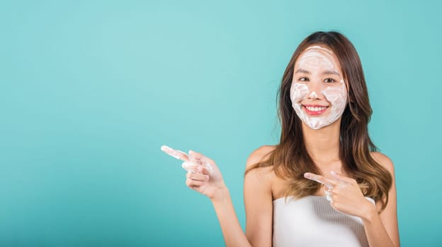 Portrait Asian young woman washing face foaming facewash soap scrub on skin studio shot isolated on blue background, female applying bubble facial foam and cleansing and pointing finger to side away