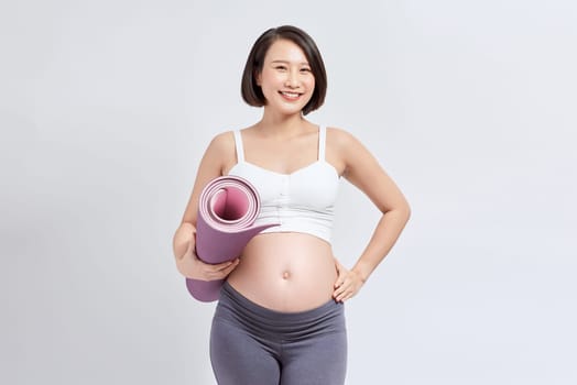 Pregnant girl holding yoga mat standing over white background.