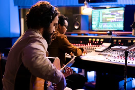 Songwriter creating a new song with his guitar in control room, playing electro acoustic instrument and singing next to record producer. Young artist performing live, editing music on mixing console.