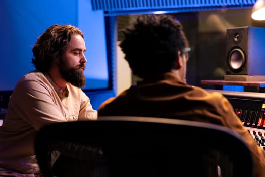 Musician songwriter composing a song with electro acoustic guitar, performing live in control room at studio. Artist collaborating with sound designer producer to create music for his album.