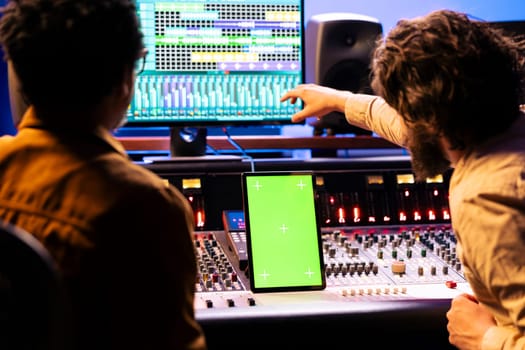 Singer and producer collaborating on new music for an album, looks at greenscreen on tablet next to control panel board in studio. Expert and musician recording new tracks, mixing console.