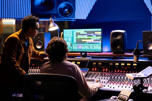 Singer and producer work on soundboard controller at recording studio station. Sound engineer uses digital console for music production, operating on faders and key buttons at control desk.