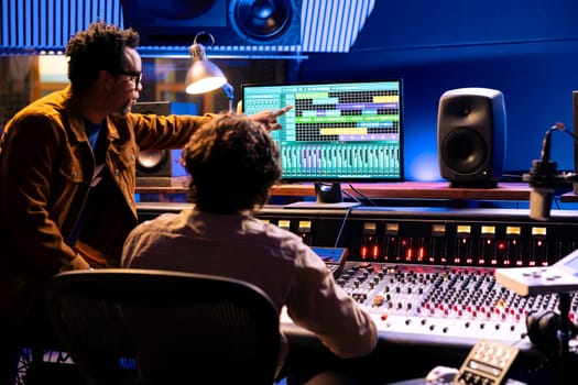 Team of artists collaborating on processing recorded files with audio software, pushing faders to adjust volume levels. Technician and singer creating music on control panel board in studio.