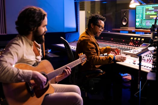 Composer playing a new hit song on his guitar in studio control room, recording and producing music with audio expert. Musician performing tracks on electro acoustic instrument, post production.