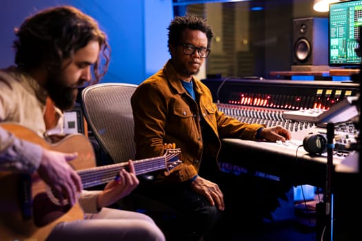 Singer recording electro acoustic guitar in professional music studio, playing musical instrument to compose a new hit song with his producer. Diverse musicians singing a record in control room.