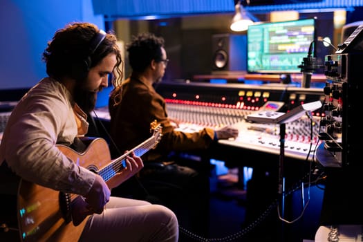 Skilled artist musician recording his guitar sounds in professional studio, using electro acoustic tunes to edit another record for an album. Singer composing music with audio expert in control room.