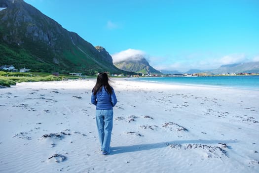 A woman walks along a pristine white sand beach in Norway, with a stunning mountain backdrop and clear blue water. Ramberg Beach Lofoten Norway