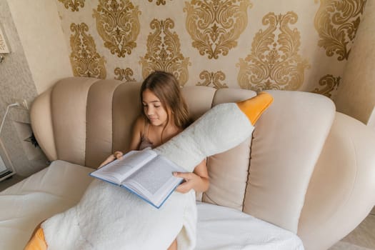 A girl is reading a book while sitting on a bed with a stuffed duck. The room has a gold and white color scheme, and there are two lamps in the room