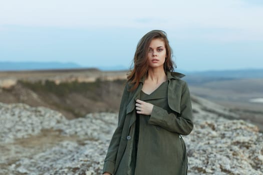 Woman in olive trench coat stands on hill overlooking vast ocean expanse