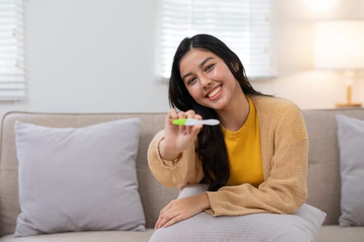 Smiling woman holding a positive pregnancy test while sitting on a comfortable sofa in a bright and cozy living room.