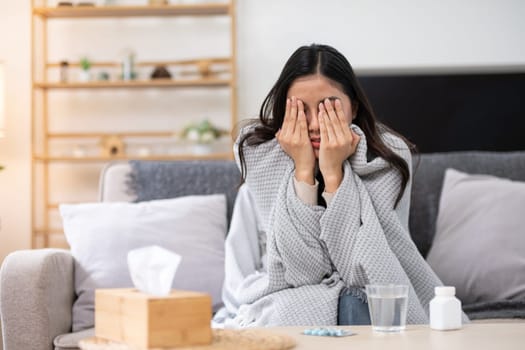 A woman sitting on a couch, wrapped in a blanket, covering her face with her hands, with medicine and tissues nearby, indicating illness or discomfort.