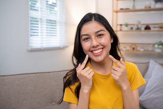 A cheerful woman in a yellow shirt smiles and points at her cheeks while sitting on a couch in a cozy living room.