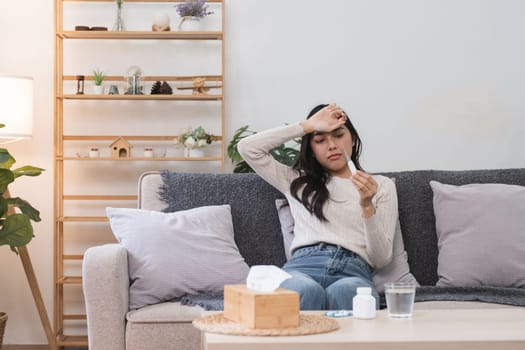 A young woman sits on a couch, checking her temperature with a thermometer, surrounded by home decor and health items.