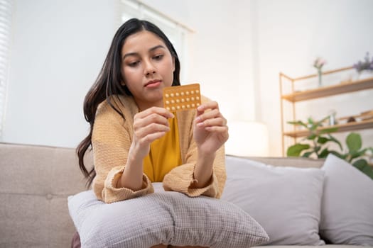 A young woman examines a pack of birth control pills while sitting on a couch in a modern living room setting.