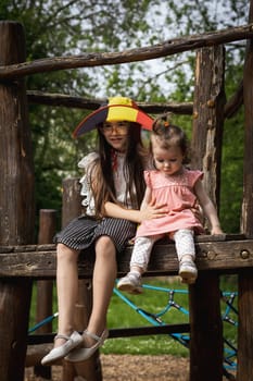 Portrait of two beautiful Caucasian brunette little girls in a Belgian flag hat sitting on a wooden walkway of a rope swing in a city park on a summer day and looking to the side, close-up side view.