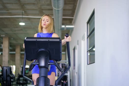 Blonde fit young caucasian woman exercising using elliptical machine in the gym