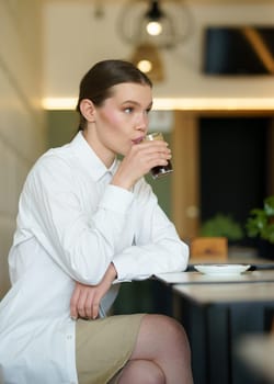 Vertical three quarter length photo of a sensual and elegant caucasian adult businesswoman sipping coffee in a modern cafeteria at night