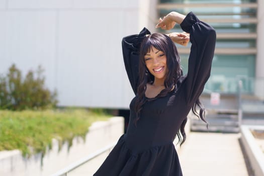 Energetic caucasuan young woman smiling at camera while dancing in the city wearing summer black dress