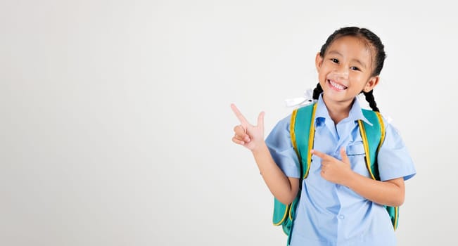 Portrait smiling Asian little girl kindergarten with backpack pointing finger at empty studio shot isolated white background, happy woman kid in pigtails wearing school uniform, back to school concept