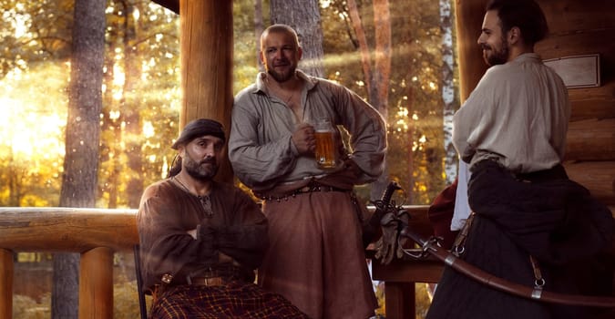 Three men in national clothes. A man in a hat is sitting, another is standing and holding a glass of beer. The third is turned with his back. Historical reconstruction. High quality photo