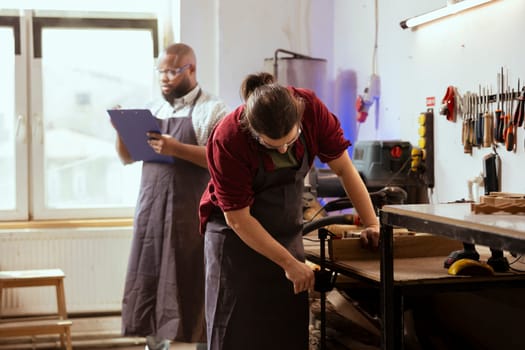 Woodworker wearing safety glasses while using bench vise to hold lumber block in workshop next to coworker. Craftsperson using protective equipment and vice tool to clamp piece of wood