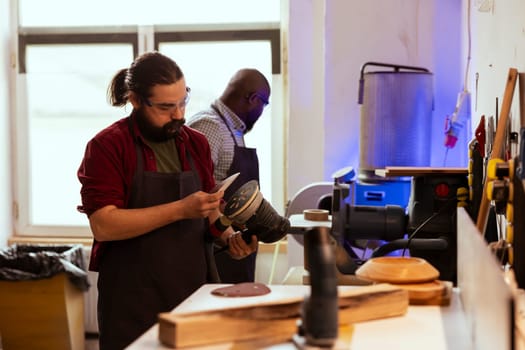 Carpenter holding power tool used to smooth surfaces by abrasion with sandpaper, changing cut off discs. Man preparing necessary equipment for furniture assembling job in studio