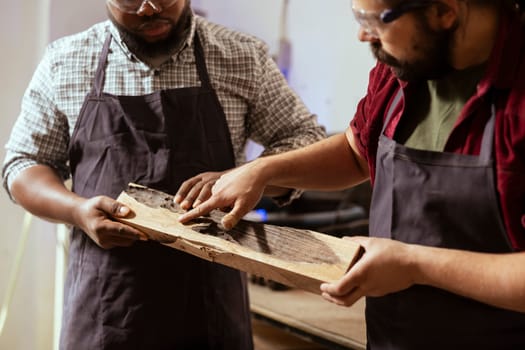 Woodworker holding timber block, showing workshop partner improvements to be made. Manufacturer pointing to apprentice defects in piece of wood to be fixed, close up shot