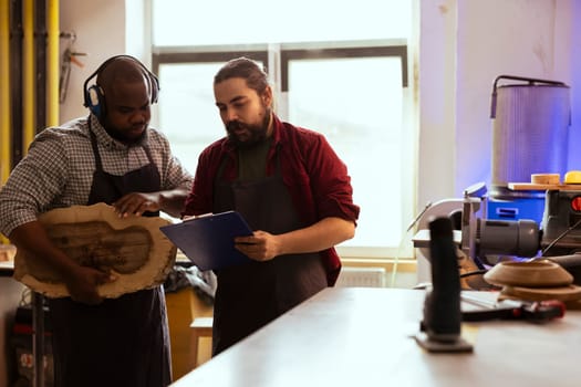 Cabinetmaker and apprentice looking over technical drawings on notepad to make creative wood art pieces. Artisan and BIPOC colleague looking at blueprints to execute woodworking projects