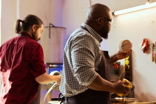 African american manufacturer doing last touches on piece of wood before using it for furniture assembly, sanding with sandpaper. BIPOC woodworking professional examining timber block for damages