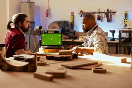 Cabinetmaker looking over blueprints schematics on isolated screen laptop in CAD software with apprentice. Woodworker and colleague designing custom 3D furniture on mockup notebook in assembly shop