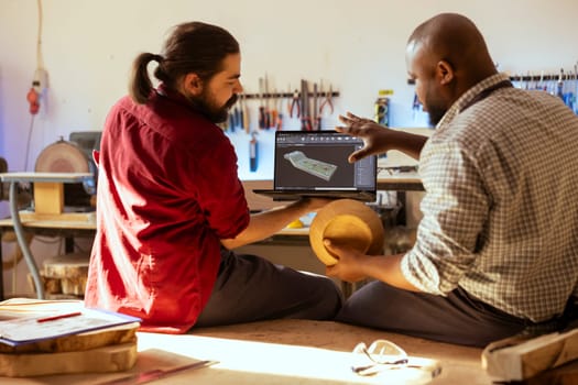 BIPOC artisan and apprentice comparing wood art piece with schematic displayed on CAD software on laptop. Woodworking experts in studio crosschecking wooden object with notebook blueprint