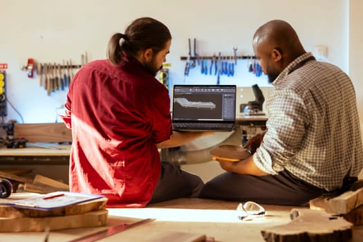 African american manufacturer and colleague comparing wood art piece with schematic displayed on CAD software on laptop. Woodworking professionals crosschecking wooden object with blueprint