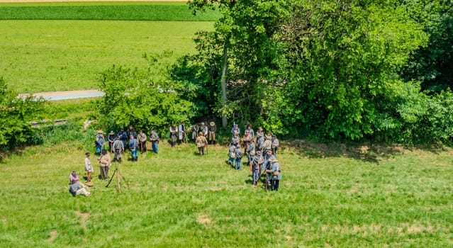 Ronks, Pennsylvania, USA, June 8, 2024 - Aerial View Of Historical Reenactors In Period Costumes Gathering In A Grassy Field Near A Tree Line, Preparing For A Scene Or Activity, With Green Farmland Extending In The Background On A Clear Day.