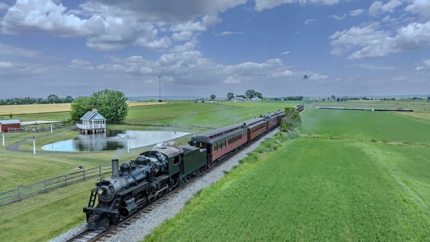 A Vintage Steam Train Travels Along A Track Through Lush Green Farmland, Passing A Pond With A Small, Picturesque House, Under A Blue Sky Dotted With Puffy Clouds In A Tranquil Rural Setting.