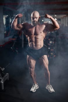 Muscular bald man posing in shorts. Bodybuilder showing off his shape in the gym