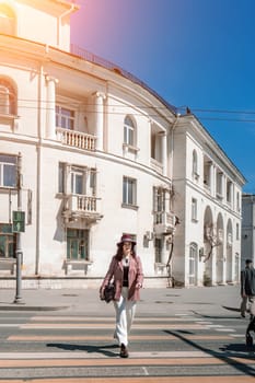 Woman city road crossing. Stylish woman in a hat crosses the road at a pedestrian crossing in the city. Dressed in white trousers and a jacket with a bag in her hands