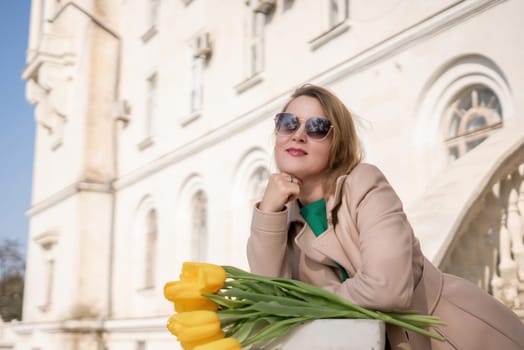 A woman wearing sunglasses and holding a bouquet of yellow flowers stands on a balcony. The scene is peaceful and serene, with the woman looking out over the city