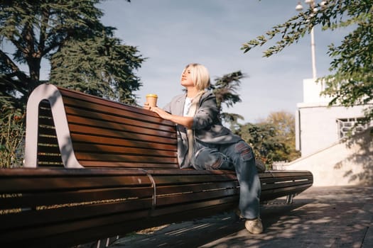 A blonde woman sits on a bench with a cup of coffee in her hand. She is wearing a gray jacket and jeans. The bench is wooden and has a curved back. The scene is peaceful and relaxing