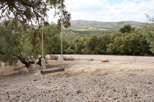 A rustic wooden swing hangs from a tree overlooking a serene countryside landscape with lush greenery, hills, and wind turbines in the background.