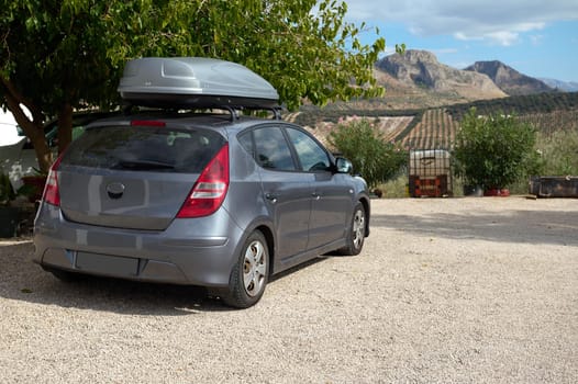 A silver car with a roof box parked on a gravel driveway with a picturesque rural landscape in the background. Perfect setting for adventure or travel themes.