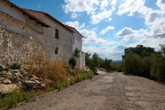 A rustic stone house in a serene countryside setting, featuring a deserted gravel road and lush greenery under a partly cloudy blue sky.