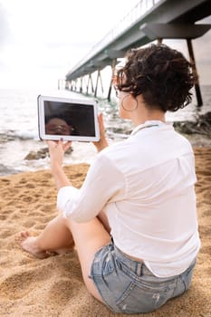 Woman on the beach with digital tablet in hand typing smiling with sunglasses