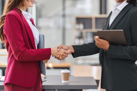Two businesswomen in formal attire shake hands with joy in a modern office, symbolizing successful partnership and collaboration.