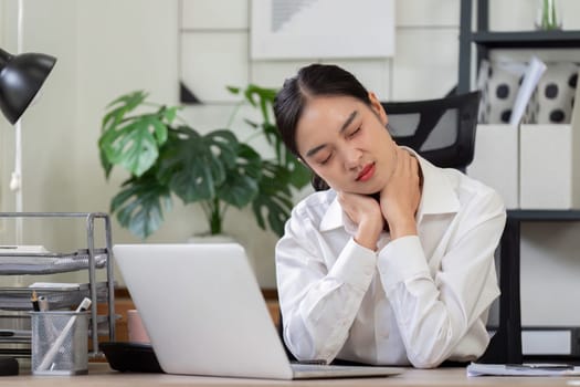 Businesswoman in white shirt suffering from neck pain while working at her desk in a modern office setting.